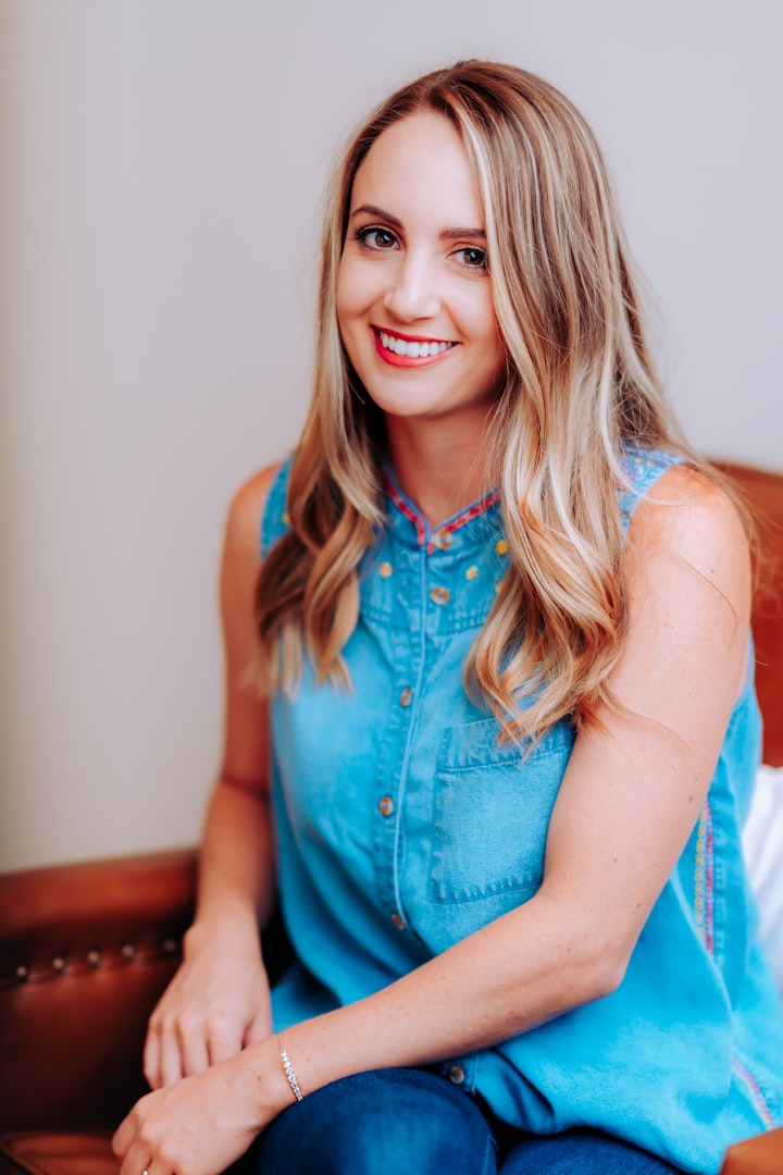 A woman with wavy, shoulder-length brown hair is smiling gently. She is wearing a white blouse and is standing in a bright, minimalist setting with turquoise decorative items in the background.